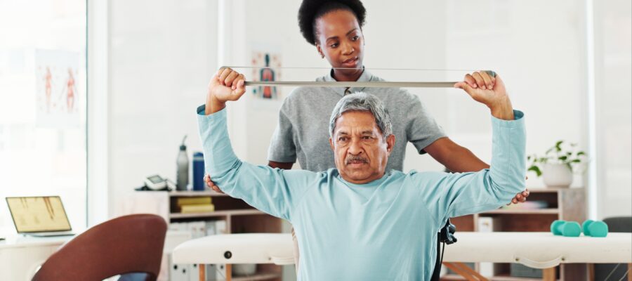 Senior man in physical therapy using resistance band with support from female therapist